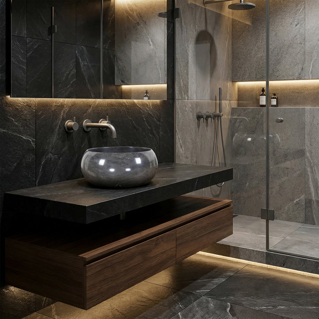 Lavatório redondo de pedra natural sobre um tampo escuro numa casa de banho moderna em estilo spa, combinado com torneira de parede, cabina de duche em vidro e iluminação indireta