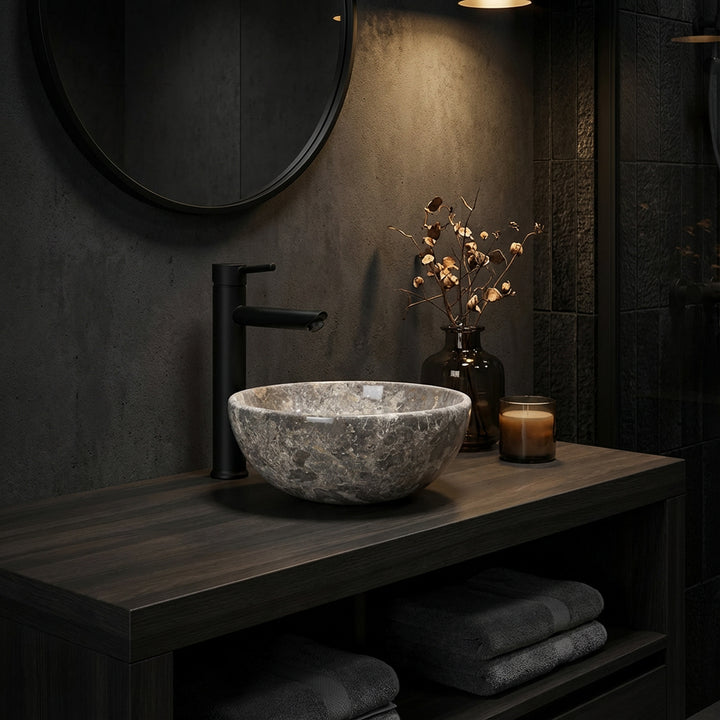 Lavatório redondo de pedra natural com veios castanho-acinzentados, assente numa consola de madeira escura, numa casa de banho com iluminação ambiente, combinado com torneiras pretas, espelho e acessórios discretos