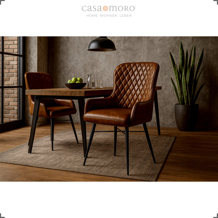 Duas cadeiras de couro castanho antigo com costura em losango, dispostas à volta de uma mesa de madeira numa sala de jantar moderna com parede de betão, piso de madeira e uma planta de interior verde.