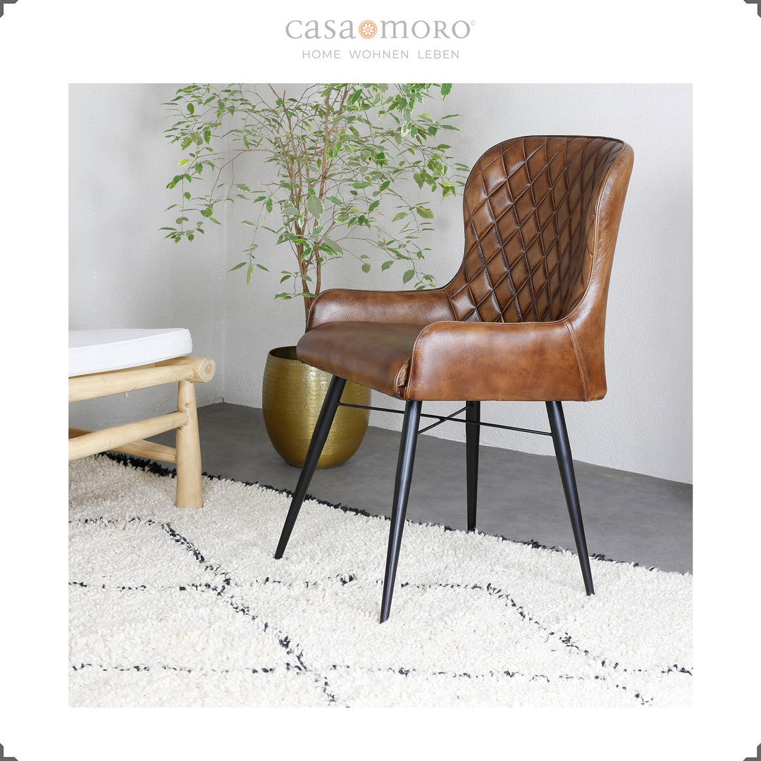 Cadeira de couro castanho antigo com costura em losango sobre um tapete claro, ao lado um vaso dourado com uma planta verde – ambiente doméstico moderno com um toque natural.