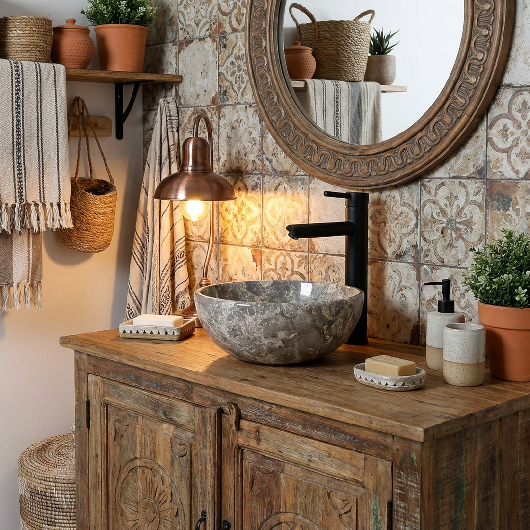 Lavatório redondo de mármore com veios cinzentos sobre um móvel de casa de banho em madeira rústica, numa casa de banho acolhedora, combinado com torneiras pretas, azulejos estampados e luz quente
