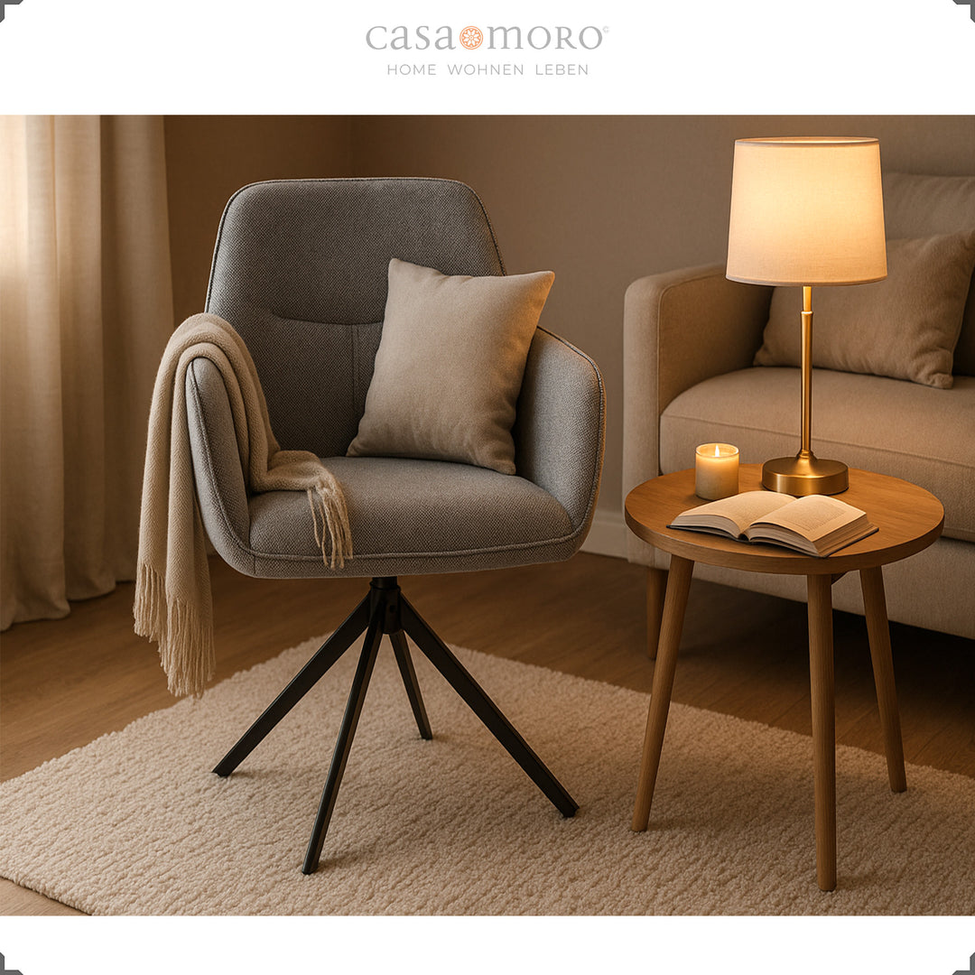 Cadeira estofada giratória a 360° com braços, em cinzento, disposta de forma acolhedora ao lado de uma mesinha auxiliar, de um candeeiro e de um sofá numa sala de estar moderna 