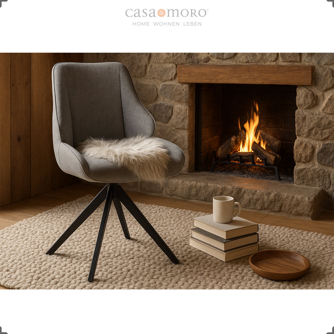 Cadeira de sala de jantar giratória a 360° em cinzento, em frente à lareira, com almofada de pele, pilha de livros e chávena – ambiente acolhedor com luz quente
