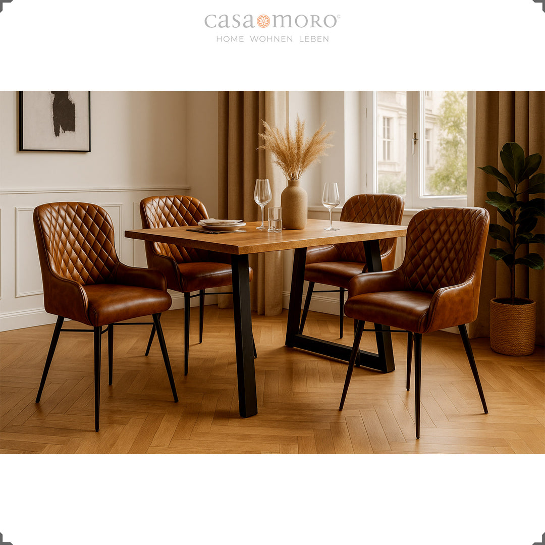 Quatro cadeiras de couro castanho antigo com costura em losango, dispostas em torno de uma mesa de madeira numa elegante sala de jantar com piso em parquet, revestimento de parede claro e decoração natural.