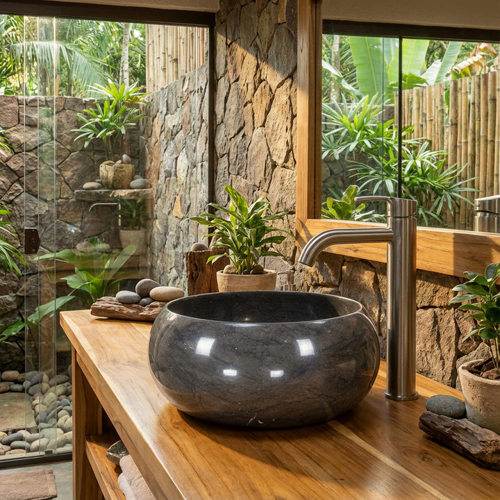 Lavatório redondo em pedra natural sobre um móvel de madeira, numa casa de banho tipo spa em plano aberto, com parede de pedra, plantas e muita luz natural