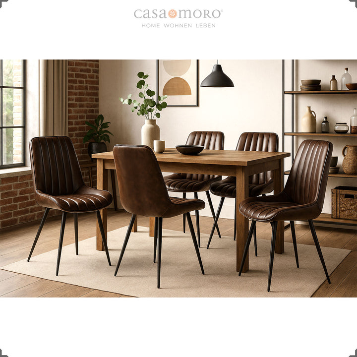 Cadeiras de couro castanho antigo com pernas de metal lacadas a preto, dispostas em torno de uma mesa retangular de madeira numa sala de jantar moderna com parede de tijolo