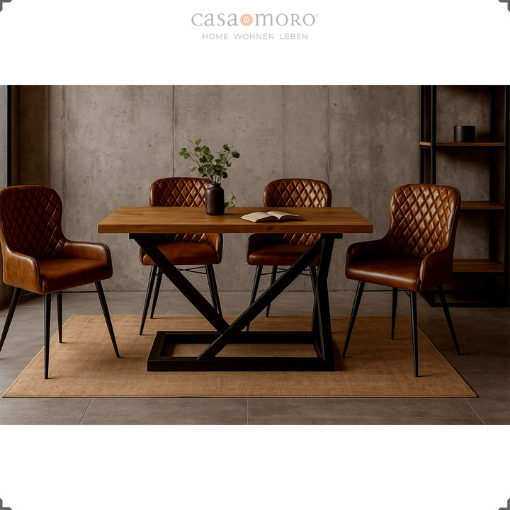 Quatro cadeiras de couro castanho antigo com costura em losango, dispostas em torno de uma mesa moderna de madeira com estrutura metálica preta, numa elegante sala de jantar em estilo loft com parede de betão.
