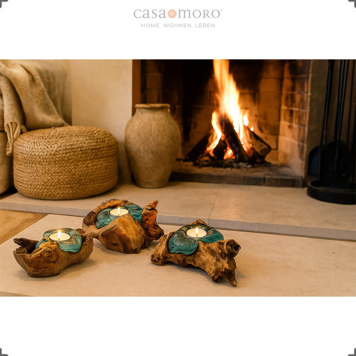 Três castiçais de vidro e madeira de raiz diante de uma lareira acesa; a sua luz suave realça o ambiente acolhedor da sala.