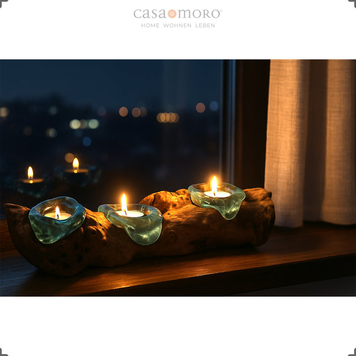 Um porta-velas alongado de vidro e madeira de raiz, com três compartimentos, colocado no parapeito de uma janela; a sua luz quente ilumina suavemente o ambiente noturno