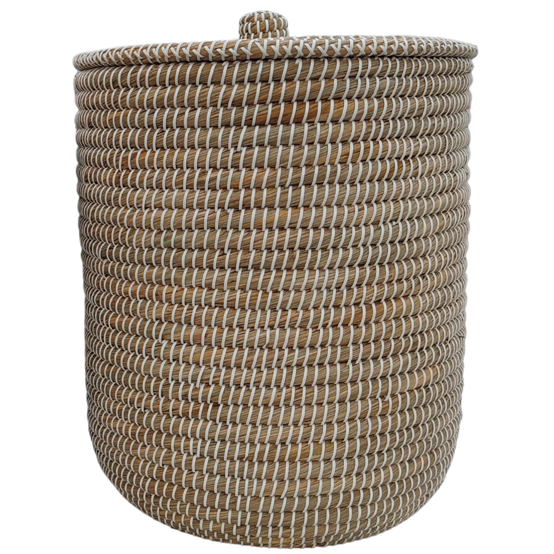 Cesto de roupa suja redondo, em erva-do-mar, na cor natural/branca, com tampa fechada e um volume de aproximadamente 65 litros.