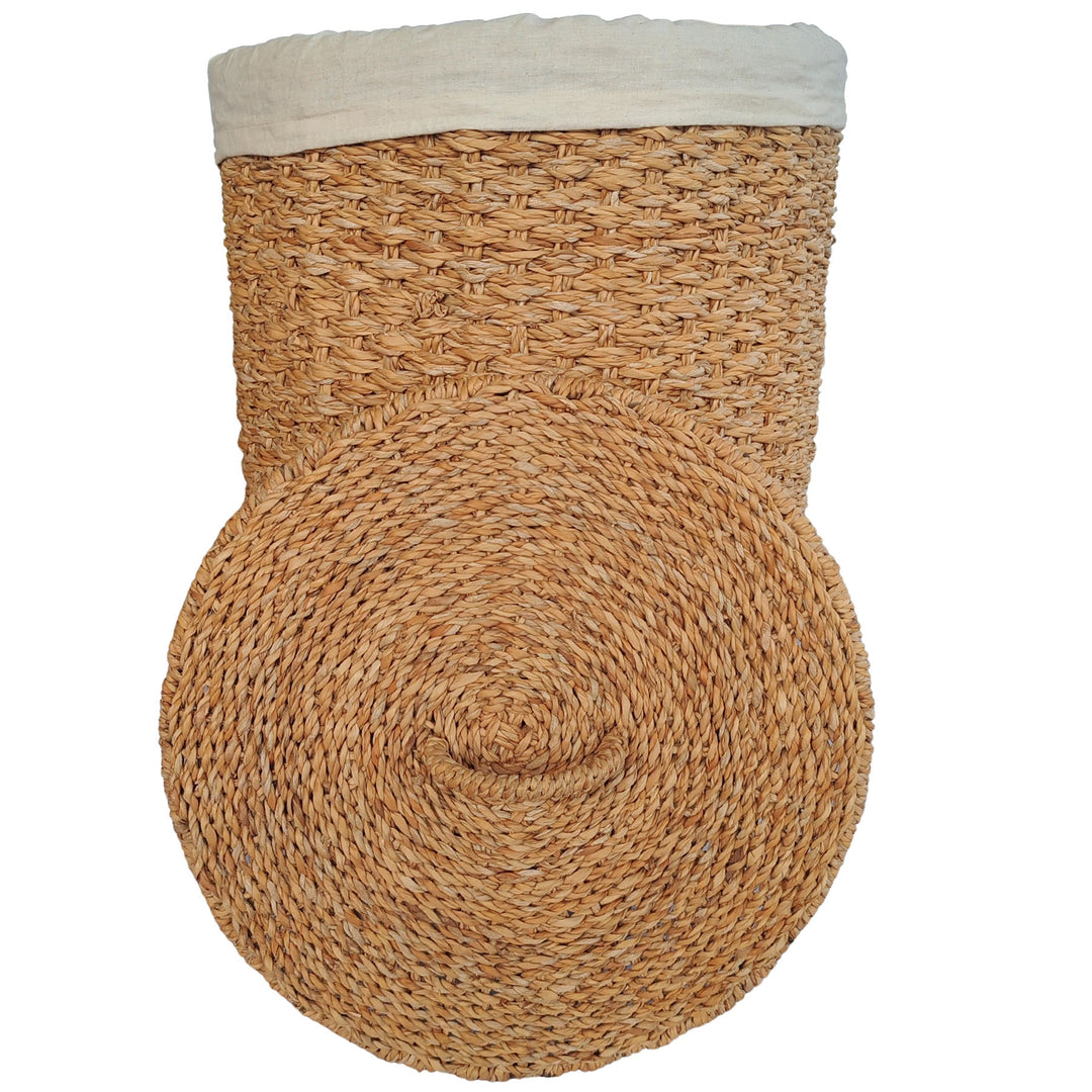 Cesto da roupa suja em erva-do-mar de cor natural, com um inserção em tecido claro e uma tampa colocada à sua frente.