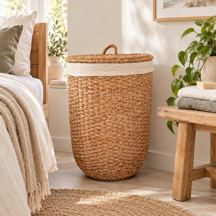 Cesto da roupa suja com tampa, feito de erva-do-mar de cor natural e com um revestimento em tecido claro, situado entre a cama e o banco de madeira no quarto.