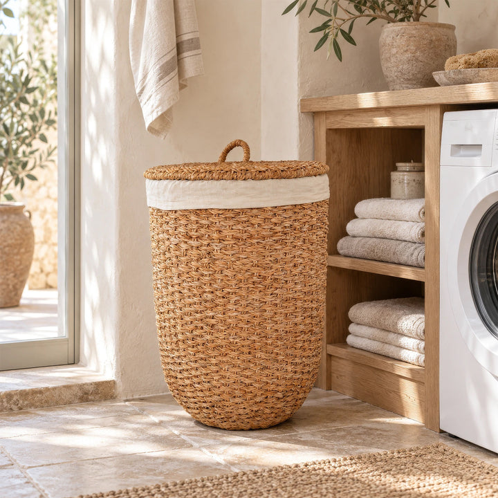 Cesto da roupa suja com tampa, feito de erva-do-mar de cor natural, junto a um suporte para toalhas e a uma máquina de lavar roupa na lavandaria.