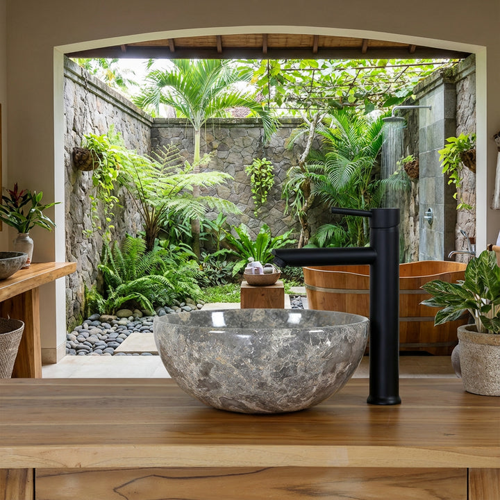Lavatório redondo em pedra natural com veios castanho-acinzentados sobre um móvel de lavatório em madeira, numa casa de banho tipo spa aberta com vista para um pátio interior verdejante, combinado com uma torneira de pé preta, uma parede em pedra natural e uma banheira de madeira
