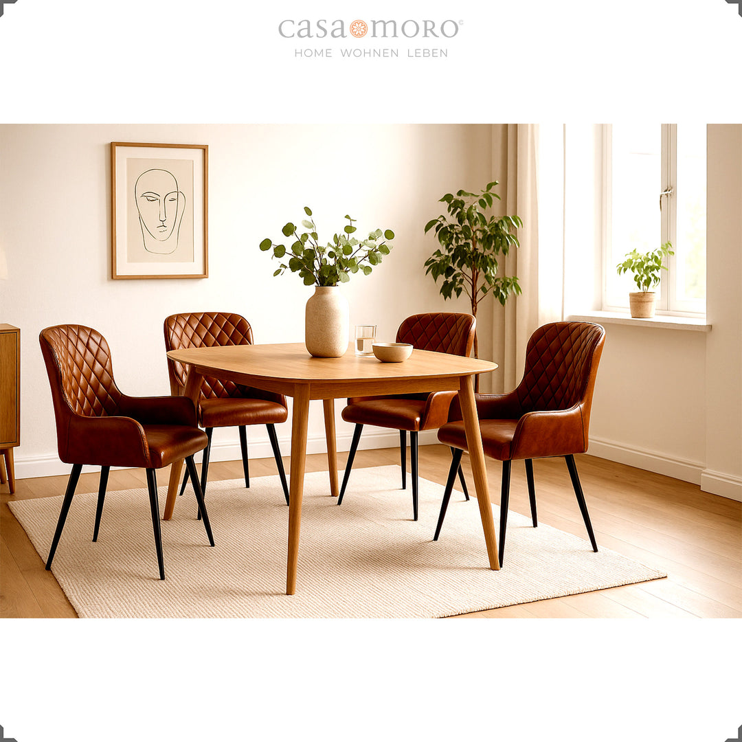 Quatro cadeiras de couro castanho antigo com costura em losango, dispostas em torno de uma mesa redonda de madeira, numa sala de estar luminosa e acolhedora, decorada com plantas e banhada por luz natural.